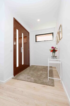 Un pasillo con una puerta de madera, una pared blanca, un ventanal pequeño y una mesa blanca con un florero.