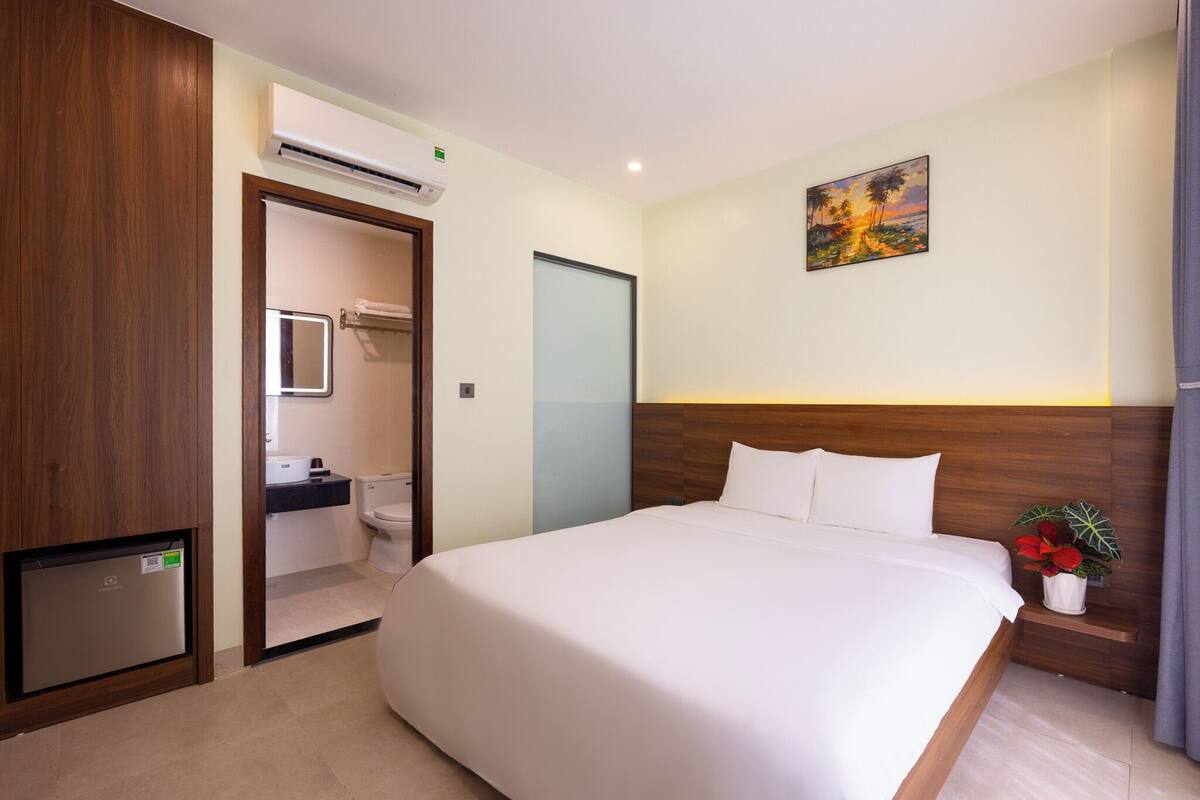 A hotel room with a large bed, a wooden headboard, a painting on the wall, a small plant on the nightstand, and an air conditioning unit.