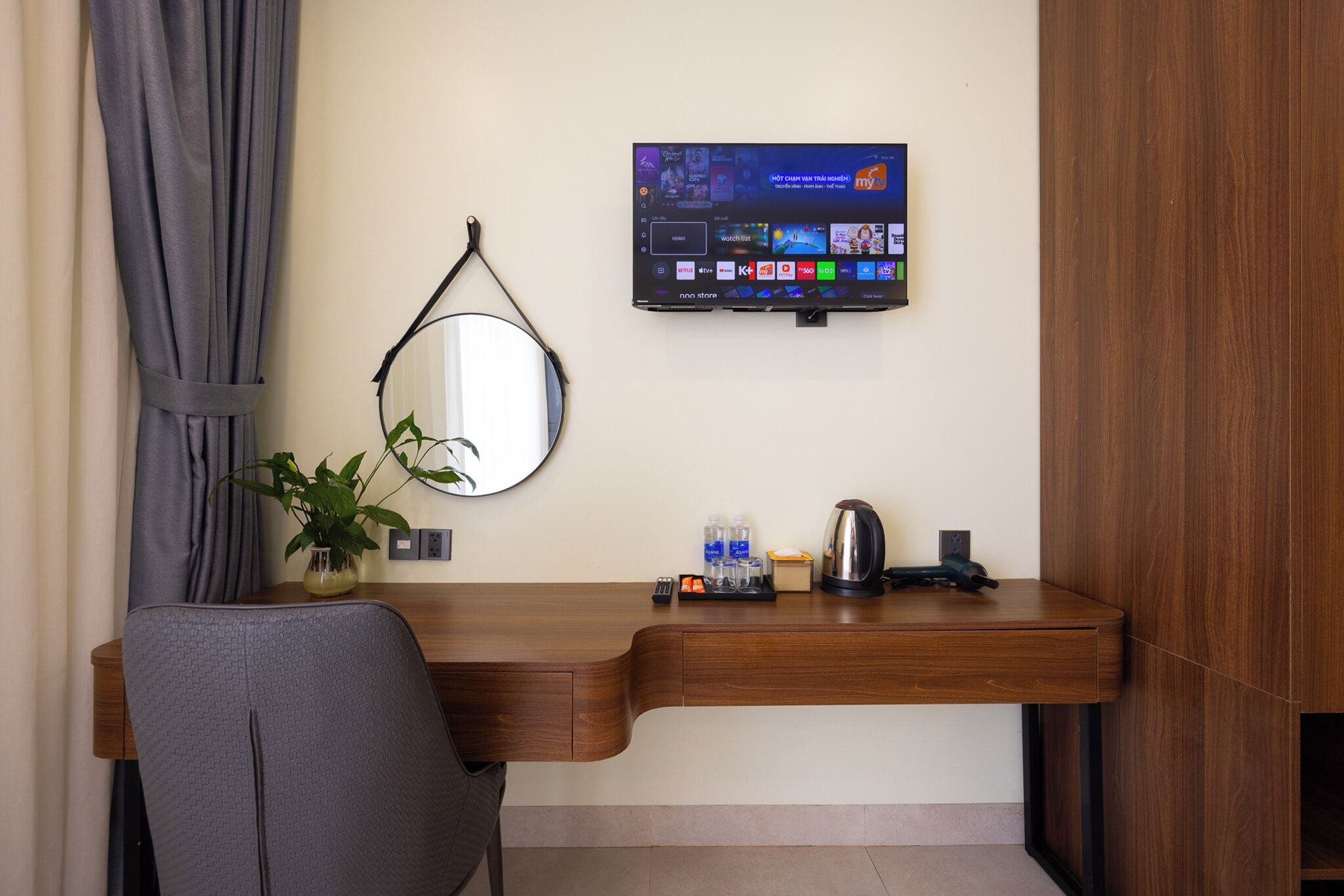 Ein Hotelzimmer mit Holzschreibtisch, flachbildfernseher an der Wand, Spiegel, Topfpflanze, Wasserkocher und Tablett mit Wasserflaschen und Fernbedienung.