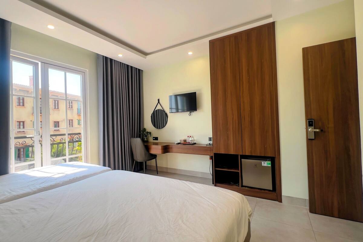 A hotel room with a bed, a desk, a television, and a view of buildings outside.