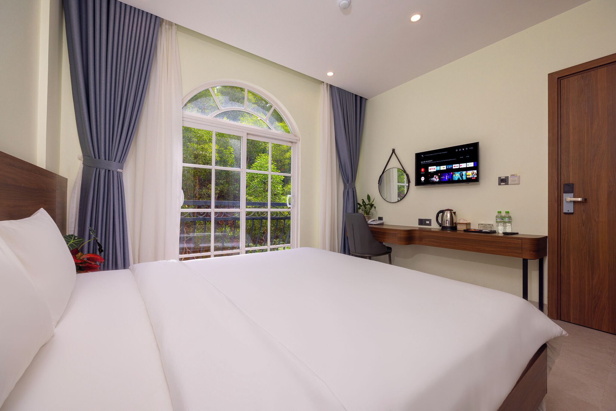 A hotel room with a large bed, a desk with a flat-screen TV, a window with curtains, and a view of greenery outside.