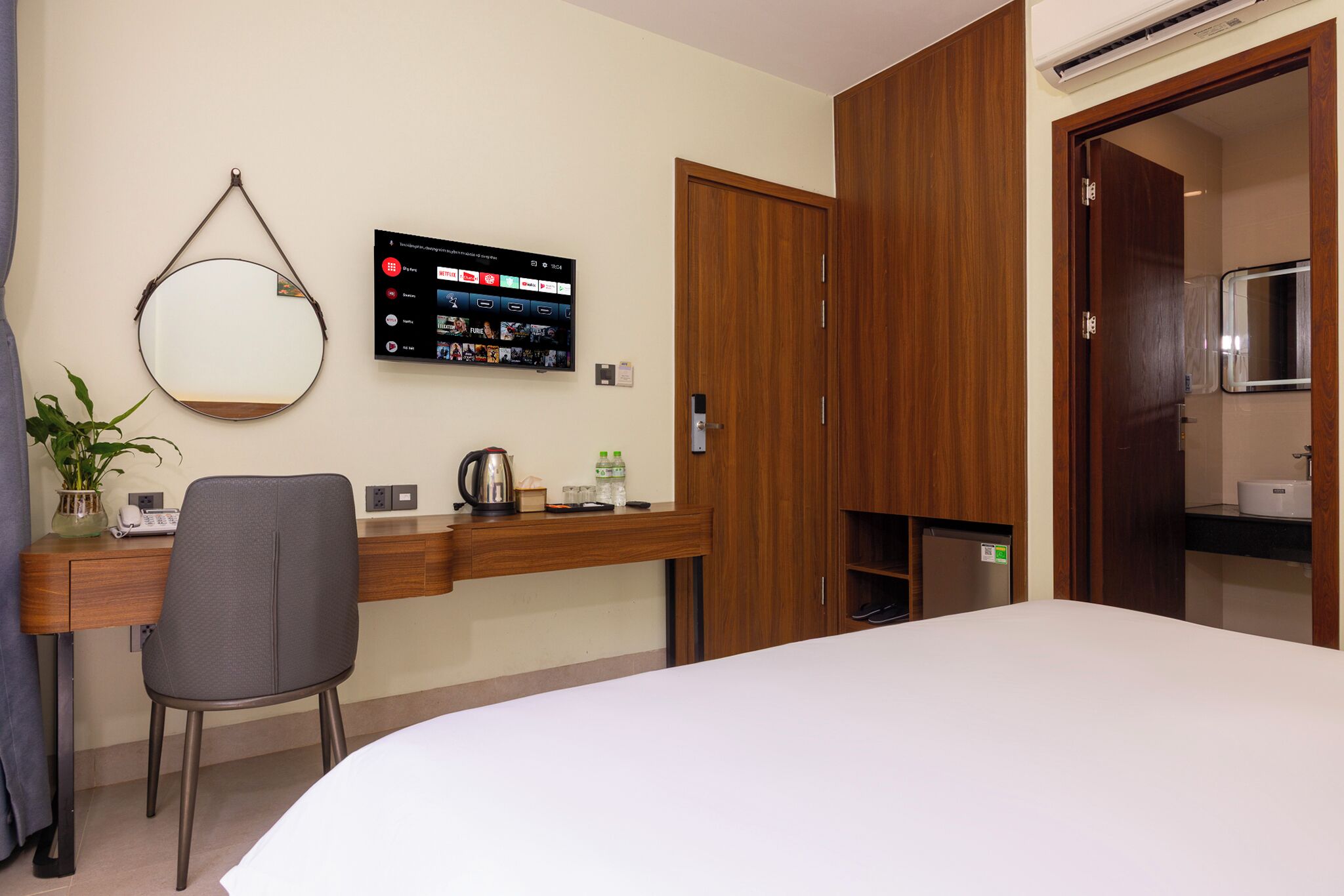 A hotel room with a bed, a desk with a chair, a TV mounted on the wall, and a wooden wardrobe.