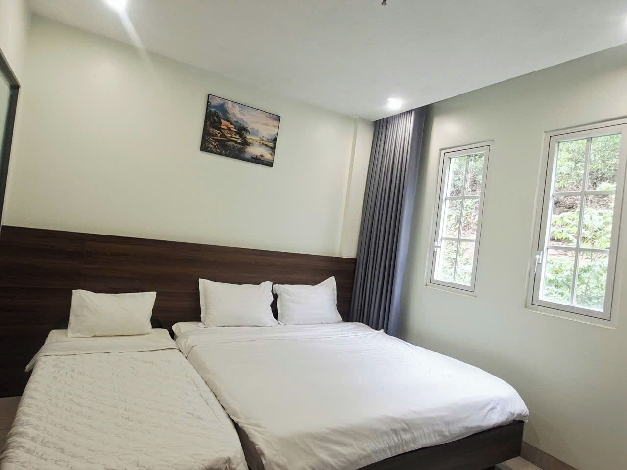 A hotel room with a large bed, a headboard, two windows, and a painting on the wall.