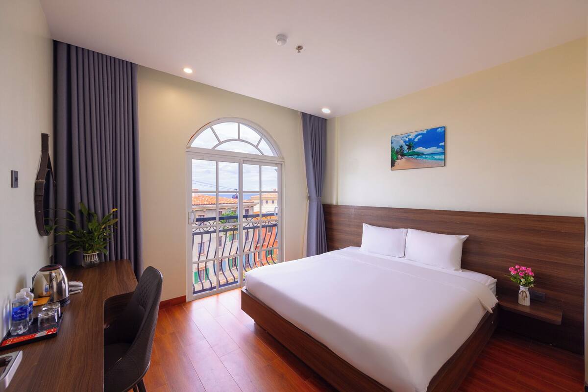 A hotel room with a large bed, a desk with a chair, a potted plant, and a view of the outside.