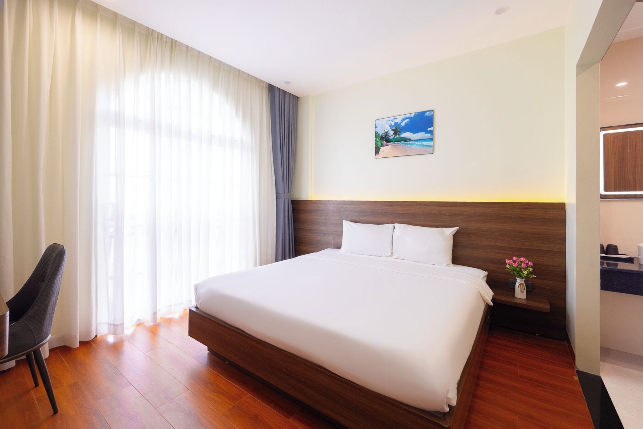 Habitación de hotel con una cama grande, cabecera de madera, mesita de noche con un florero, escritorio con una silla y un cuadro en la pared.