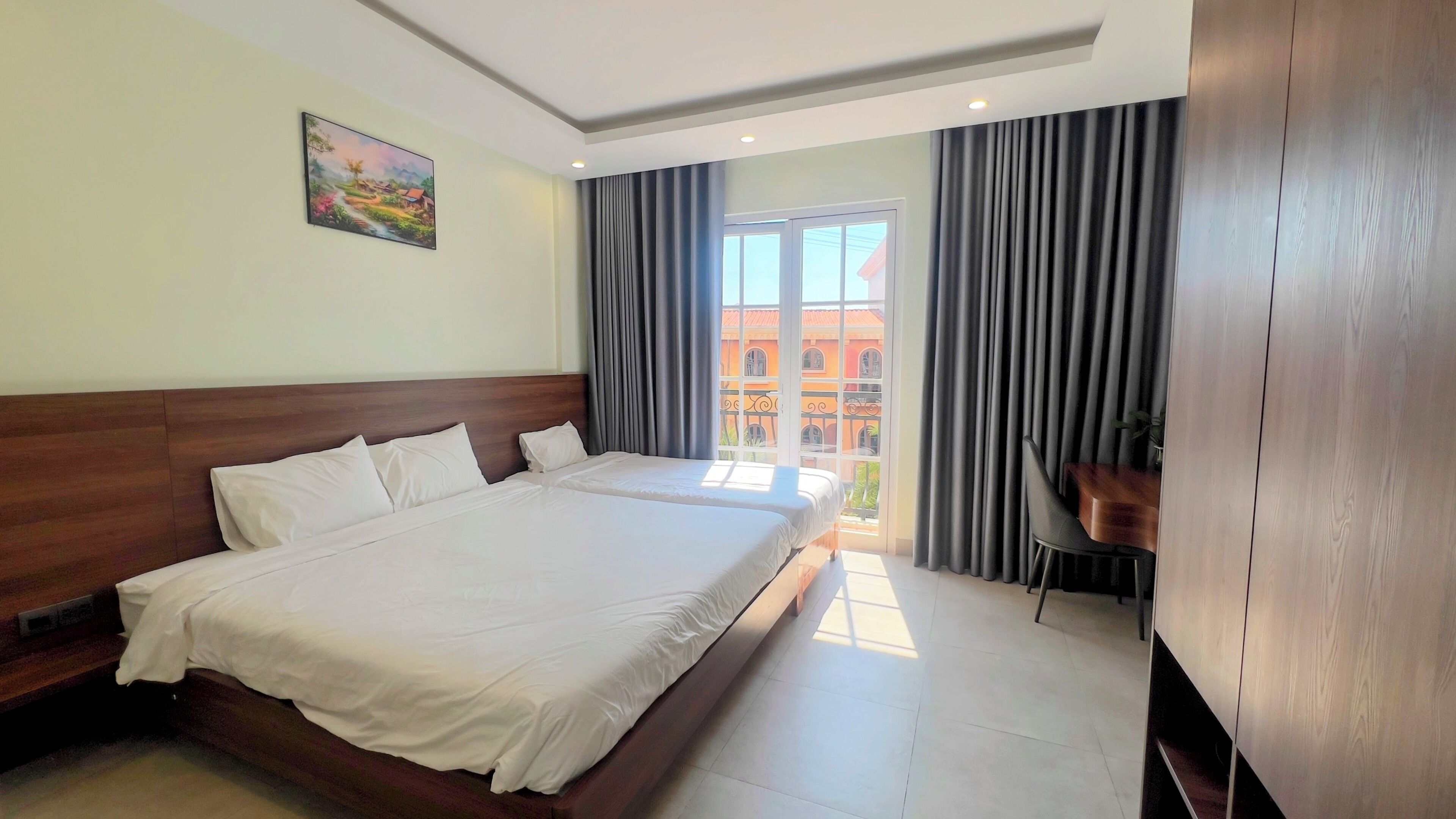 A hotel room with a large bed, a desk, a chair, and a balcony with a view of buildings.