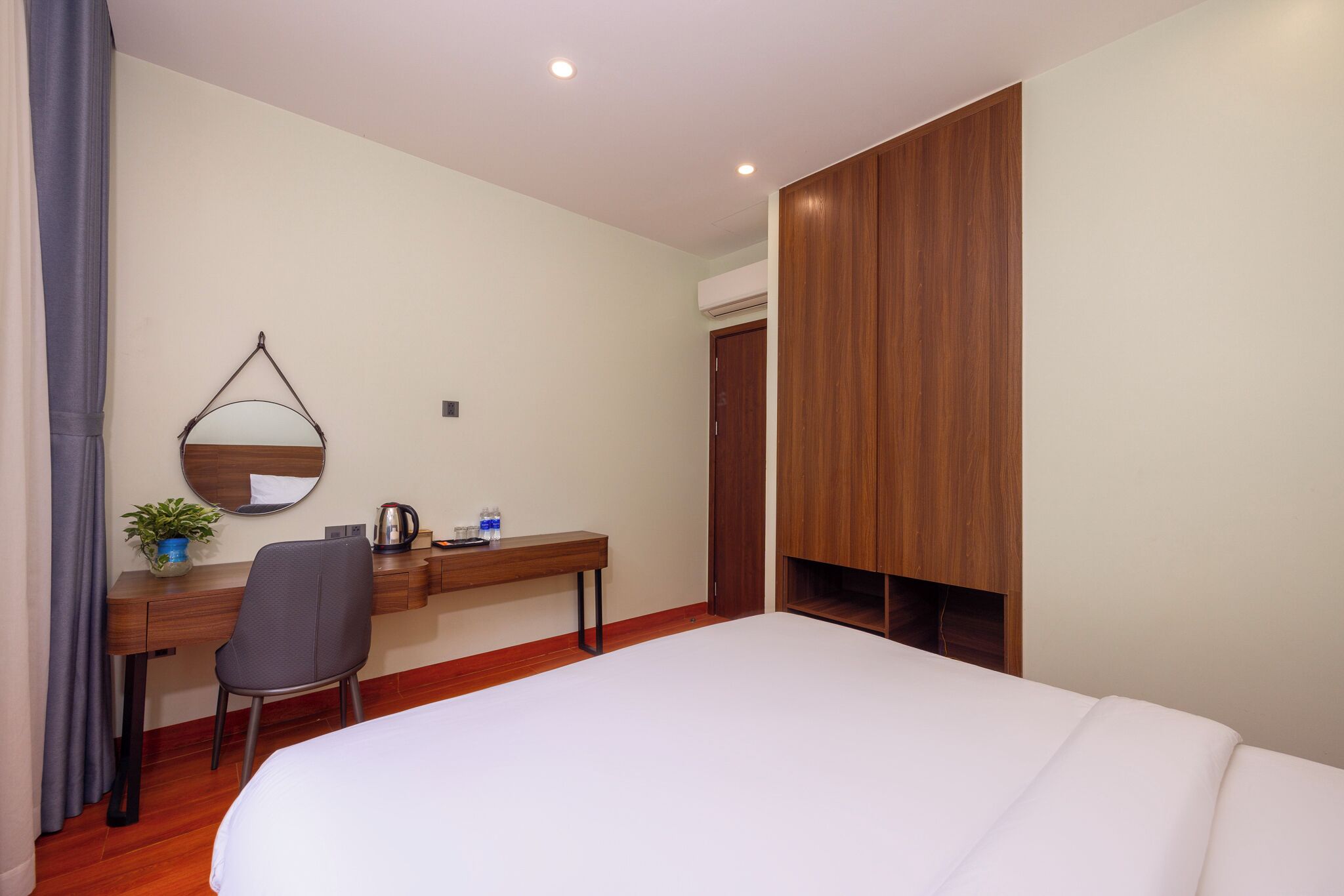 A hotel room with a bed, a desk with a chair, a mirror, a potted plant, and a wardrobe.