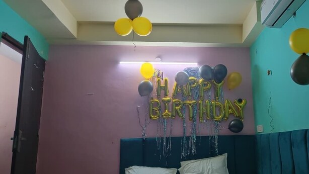 Une pièce aux murs de couleur turquoise, un lit recouvert d’une literie blanche et des ballons qui forment les mots « JOYEUX ANNIVERSAIRE ».