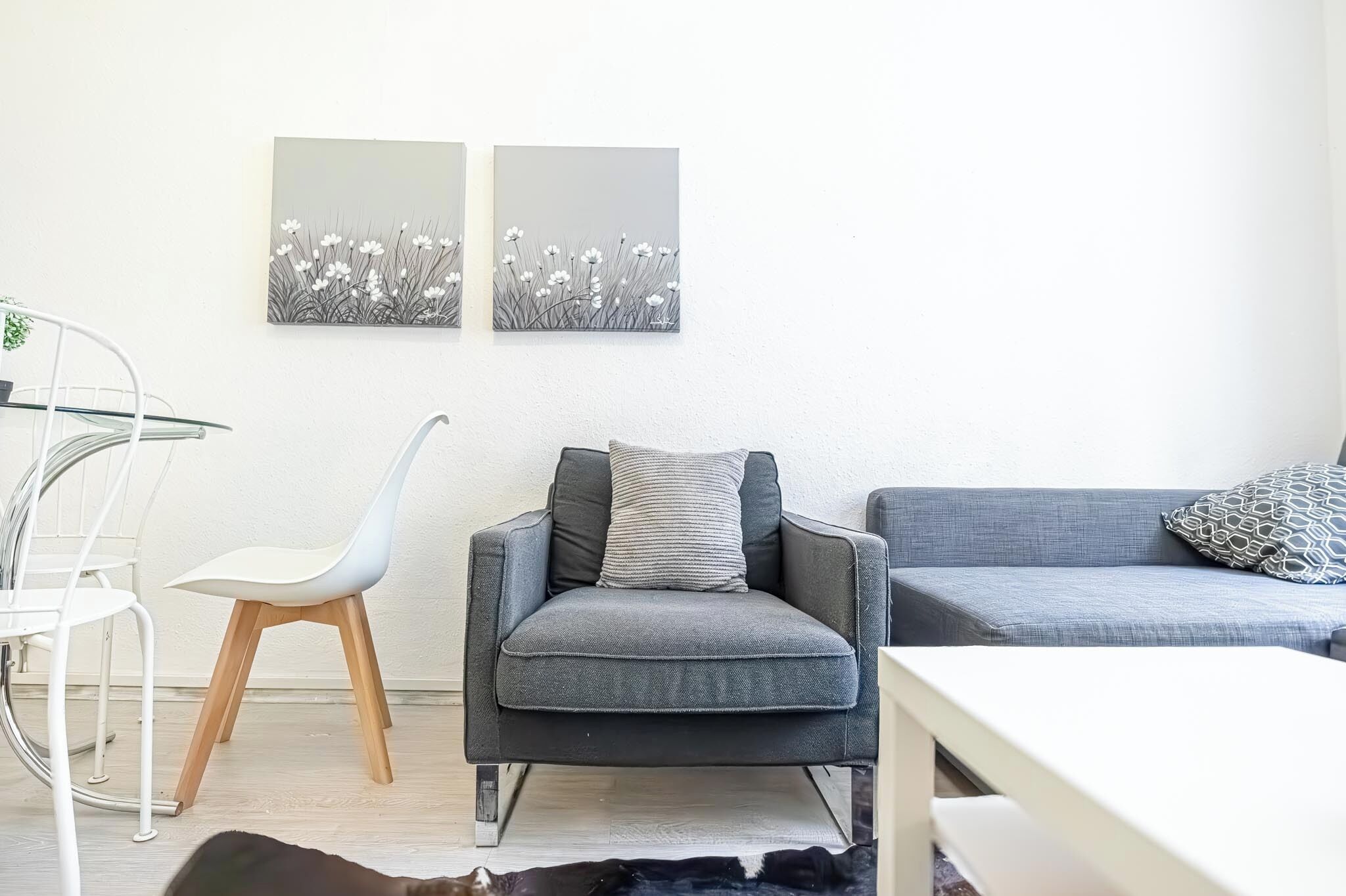 Un salon moderne avec un canapé gris, un fauteuil blanc et une table en verre. Deux œuvres d’art sont accrochées au mur et représentent des fleurs.