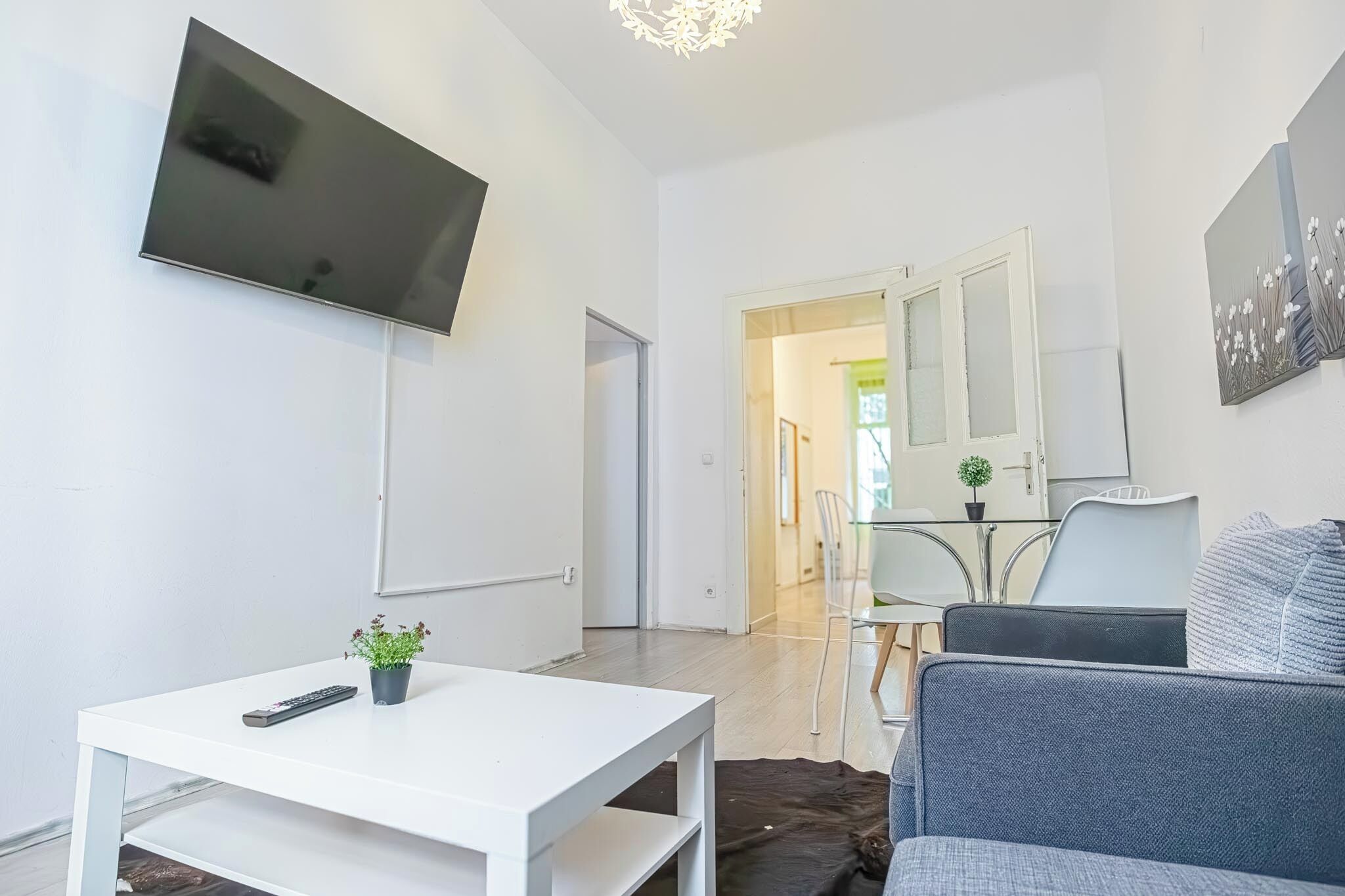 Un salon moderne avec un téléviseur à écran plat fixé au mur, une table blanche avec une télécommande et un pot de plante, un canapé gris et une table en verre avec des chaises.