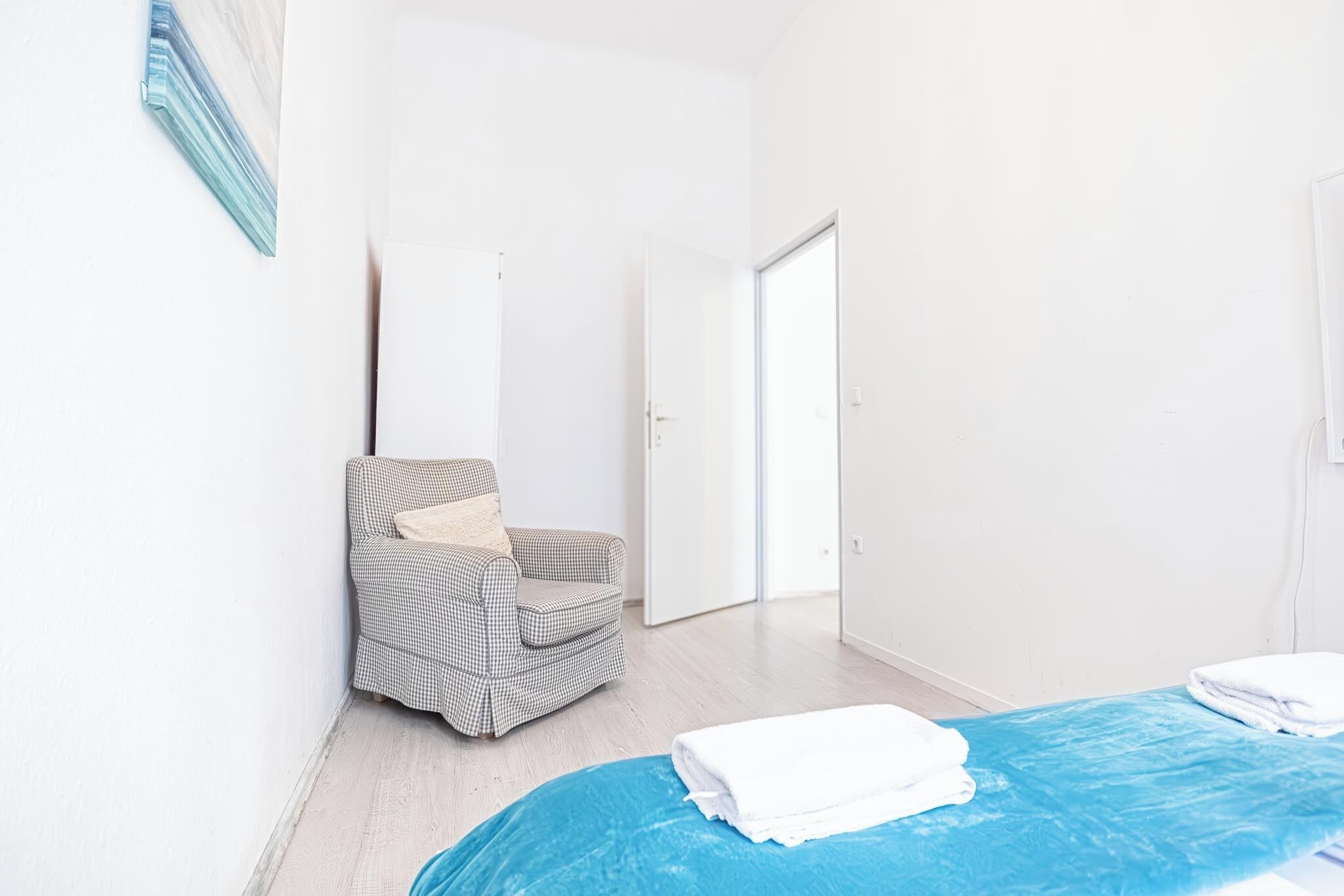 A hotel room with a checkered armchair, a blue bedspread, and a white door leading to another room.