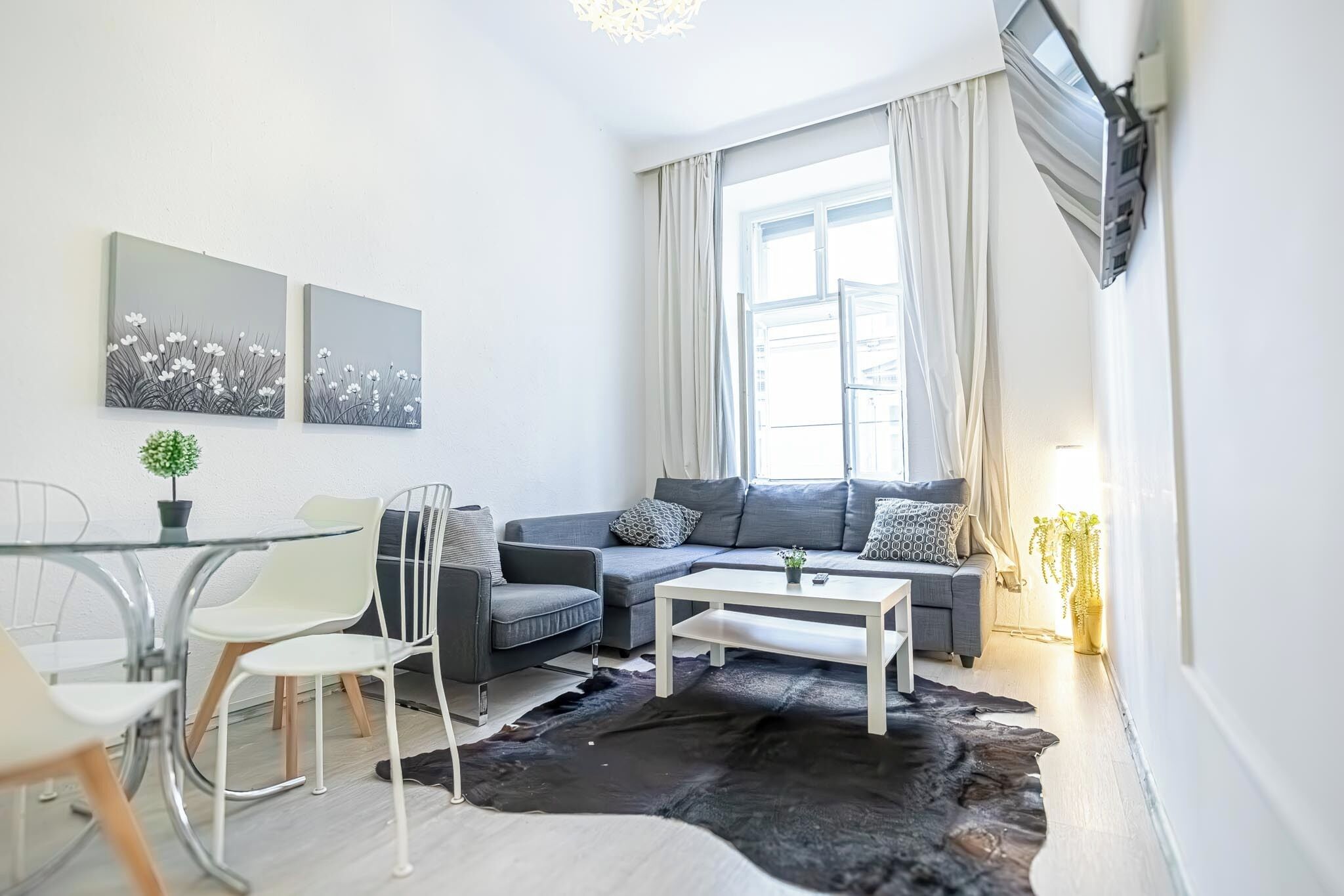 A modern living room with a grey sofa, white coffee table, and a black and white rug. There are two wall-mounted artworks and a small potted plant.
