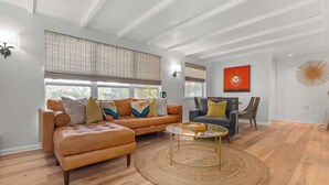 Ein Wohnzimmer mit einer Couch, einem Sessel und einem Couchtisch, großen Fenstern mit Jalousien und einer Wandlampe.
