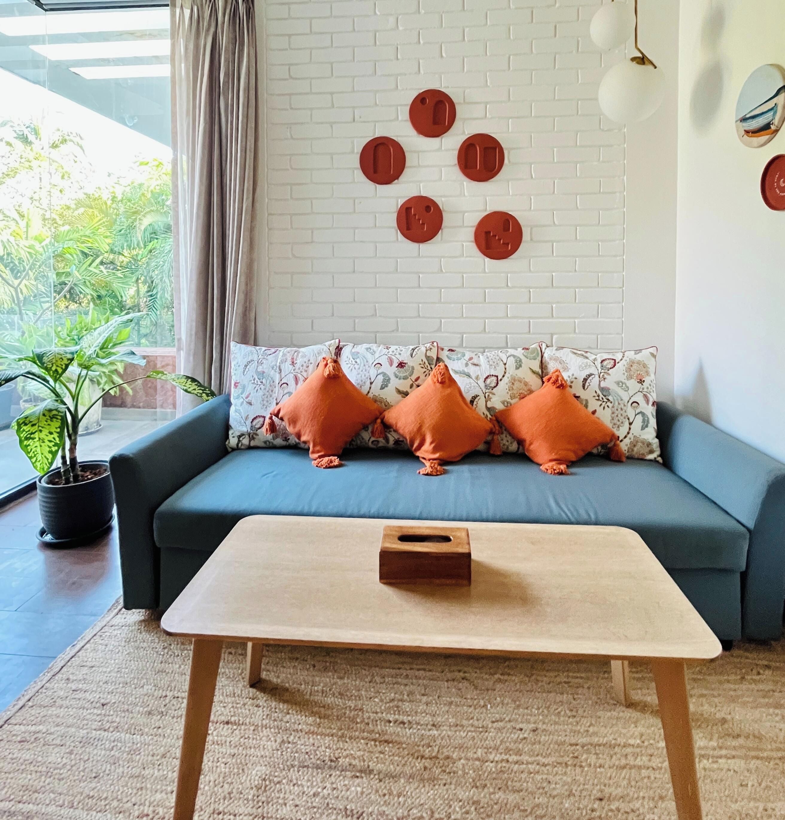 A living room with a blue sofa, orange cushions, a wooden coffee table, a potted plant, and a white brick wall with circular decorations.