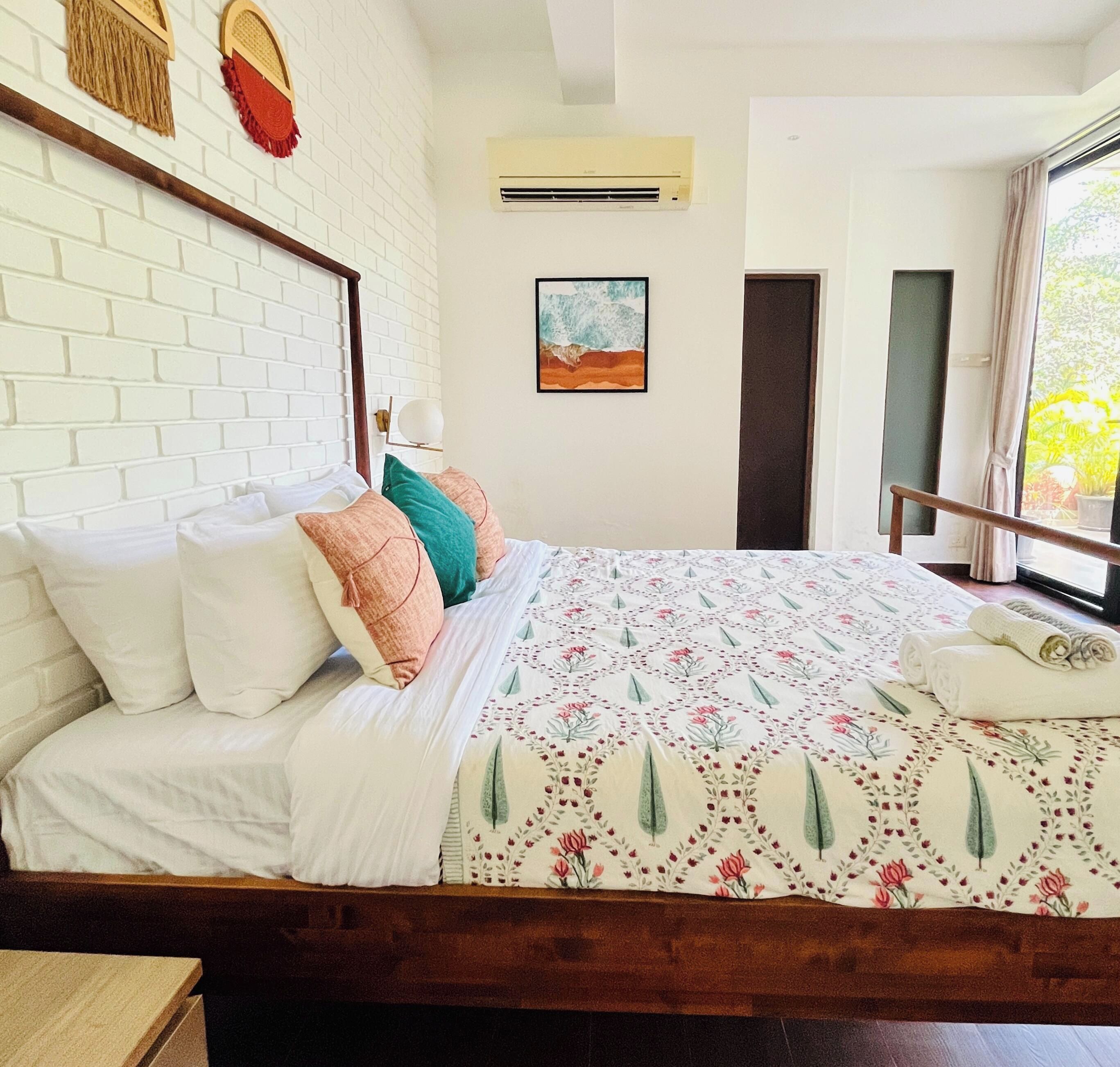 A hotel room with a white brick wall, a bed with floral bedding, a painting, and a window with a view.