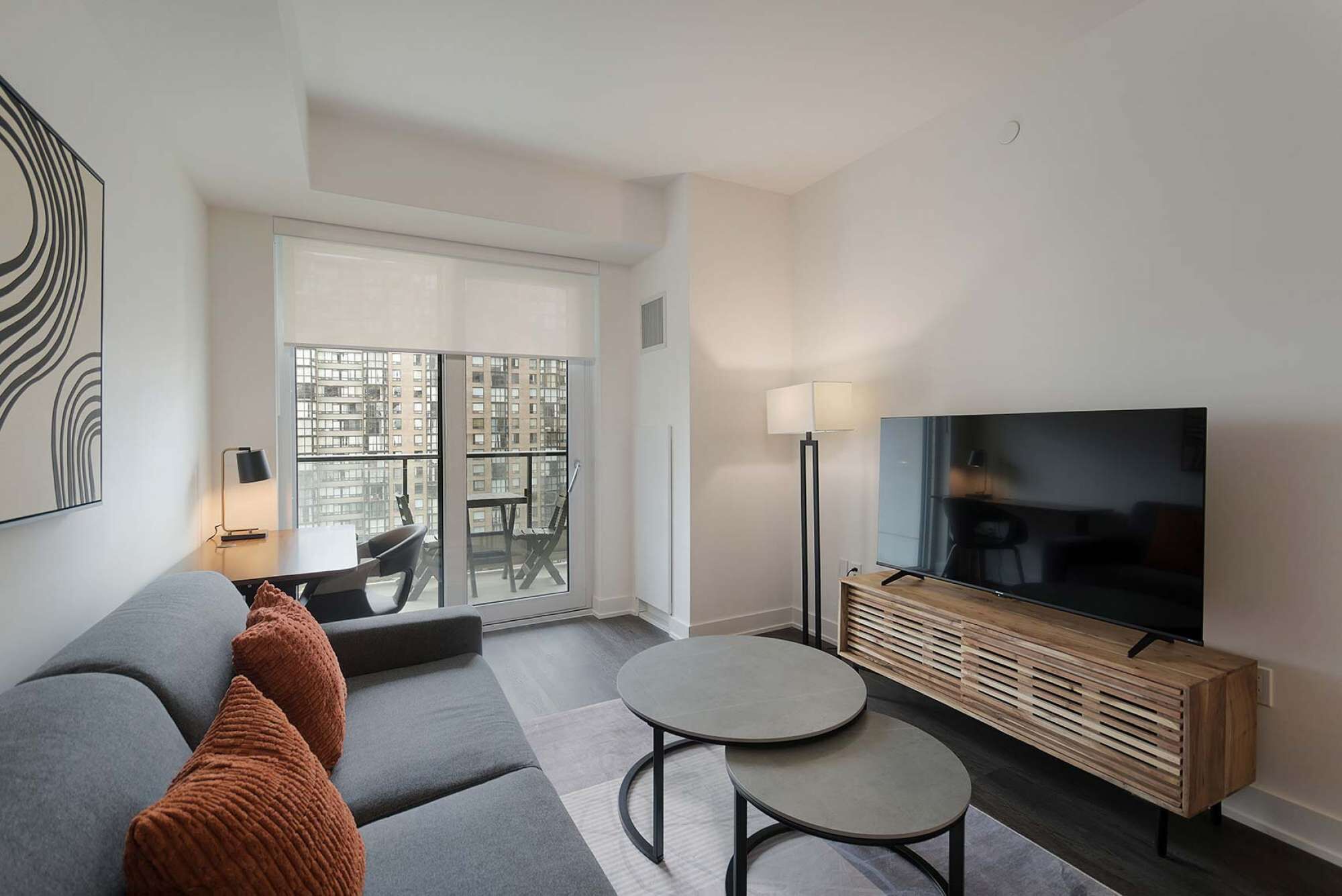 Ein modernes Wohnzimmer mit einer Couch, zwei runden Tischen, einem Fernseher und einem Balkon mit Blick auf die Stadt.