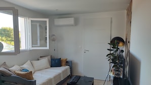 Ein kleiner, gut beleuchteter Raum mit einem Bett, einem Schreibtisch, einem Standventilator und einer Topfpflanze.