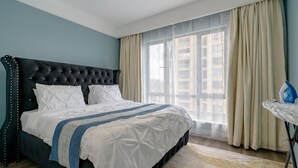 A neatly made bed with a dark headboard, a large window with sheer curtains, and a small table with an ironing board. - Alba Gardens B1102 (Nairobi)