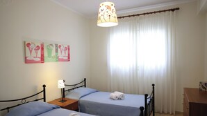A room with two beds, a nightstand with a lamp, a wooden dresser, and a window with curtains. - Entire home in Lilikas, Greece (Kalamoti)