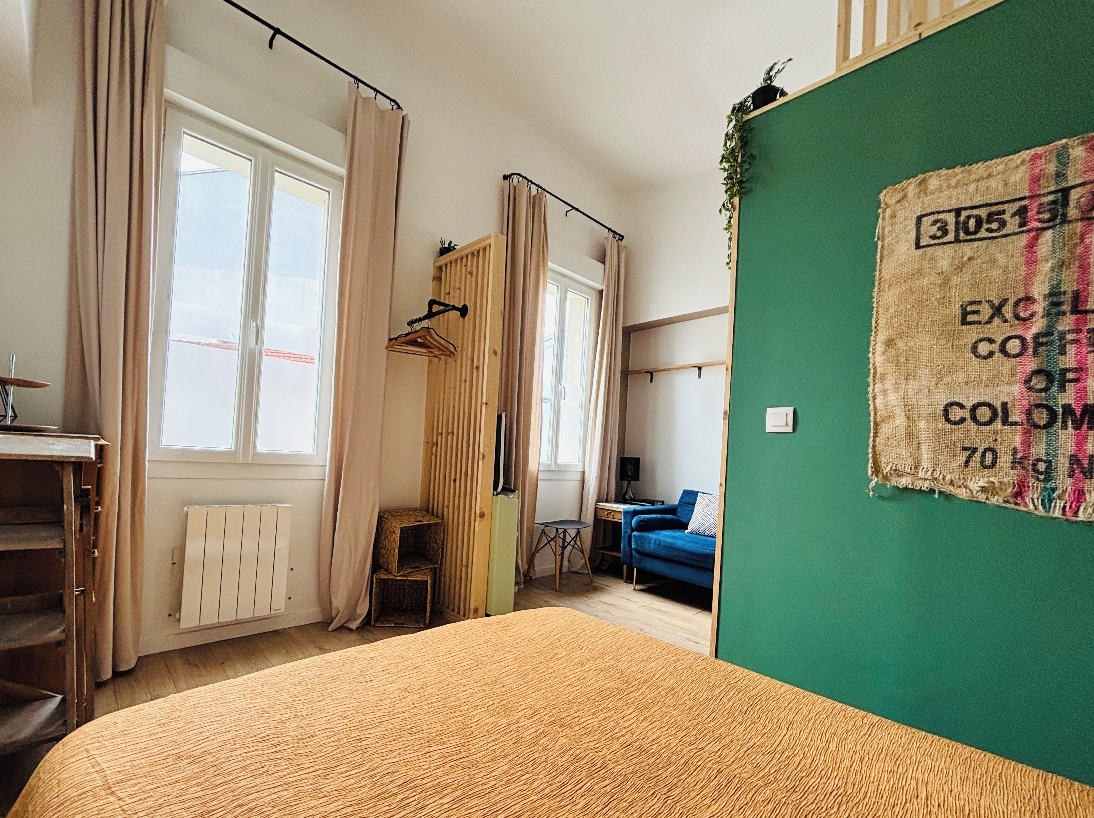 Ein Zimmer mit einem Bett, einem Heizkörper, einem Fenster mit Vorhängen, einem kleinen Tisch und einer grünen Wand mit einem Korkbrett.