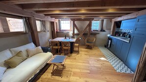 Een gezellige kamer met een houten plafond, een bank, een eettafel met stoelen, en een keukenhoek met kastjes en een spoelbak.