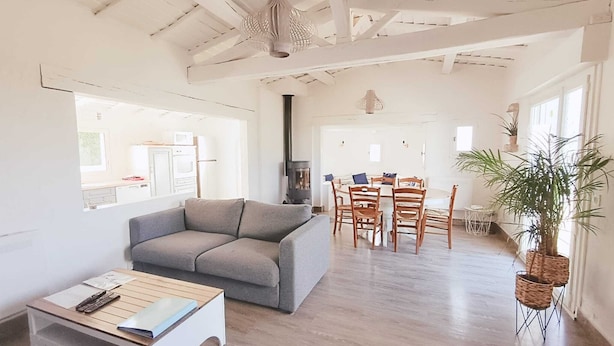 Ein geräumiger Wohnbereich mit grauem Sofa, einem Holztisch und einem Esstisch mit Stühlen. Ein großes Fenster lässt viel Tageslicht herein und bietet einen Ausblick nach draußen.