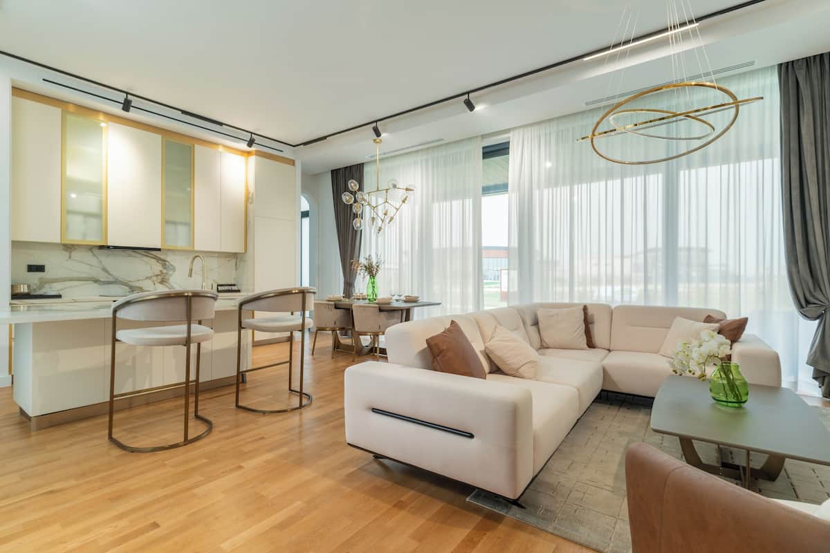 Uma sala de estar moderna com um sofá, mesa de centro e banquetas. Há uma área de cozinha com bancada de mármore e armários.