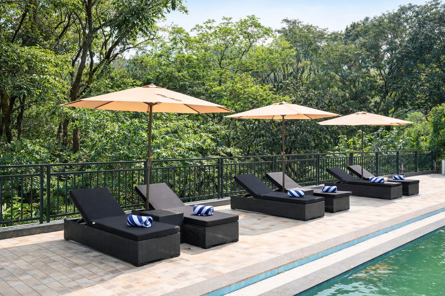 A poolside area with lounge chairs and umbrellas, surrounded by a metal railing and lush greenery.