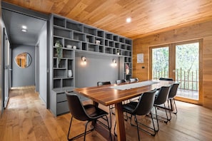 Uma zona de refeições moderna, com mesa de madeira e cadeiras pretas, rodeada por estantes cinzentas com plantas.