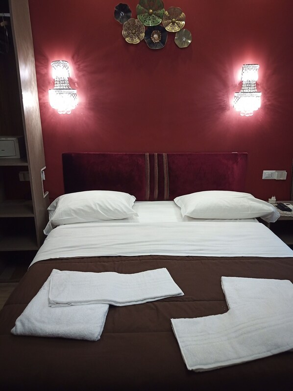 Un lit bien fait, avec du linge de lit et des serviettes blanches, un mur rouge et deux lampes fixées au mur.