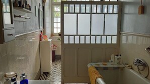 A bathroom with a bathtub, a sink, and a mirror. There are toiletries on the counter and a window with frosted glass panels. - Charming farmhouse for rent (Riachos)