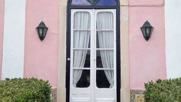 A pink building with a white door, two black wall-mounted lights, and a decorative crest above the door. - Charming farmhouse for rent (Riachos)