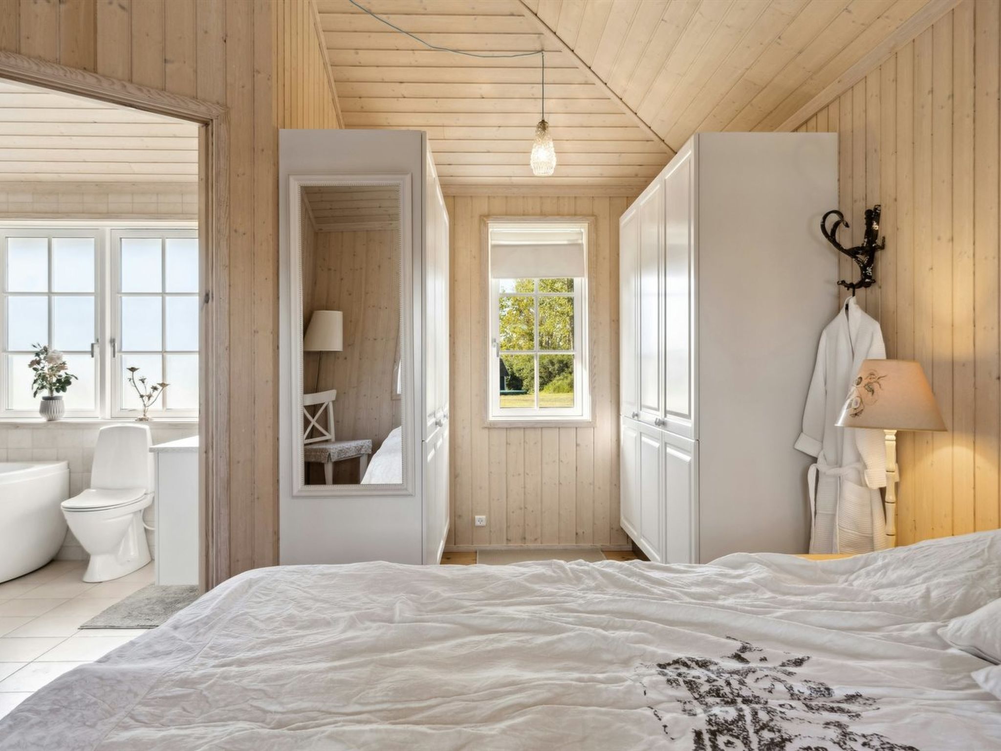 A bedroom with a large bed, a window with a view of greenery, a bathroom with a toilet and sink, and a mirror reflecting the room.