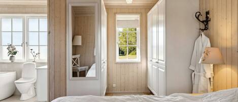 A bedroom with a large bed, a window with a view of greenery, a bathroom with a toilet and sink, and a mirror reflecting the room.