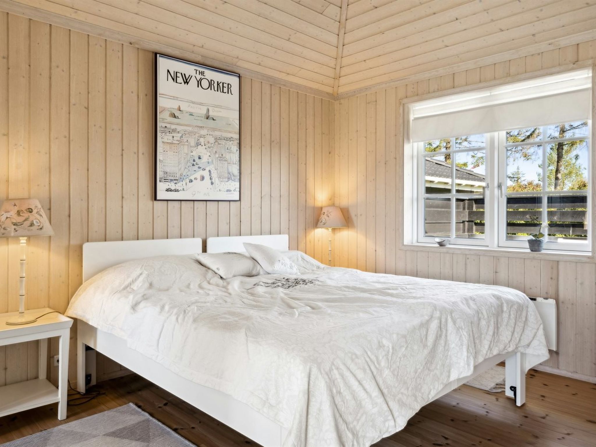 A bedroom with a bed, a nightstand, a lamp, a window with a view of trees, and a poster of New York City on the wall.