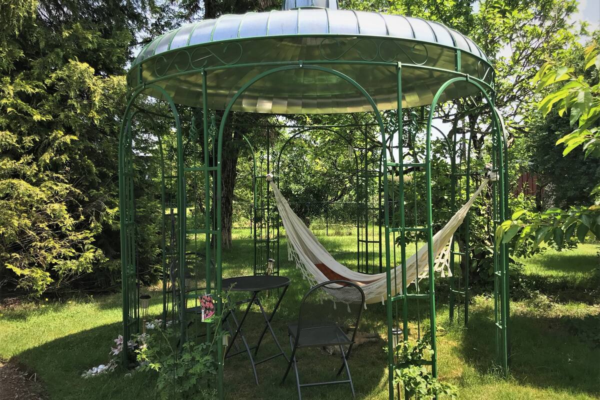 A gazebo with a hammock, surrounded by greenery and a small table with chairs.