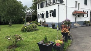 A white building with black shutters, a balcony, and a garden with various plants and flowers. - Régine MAUFFREY (Champagney)
