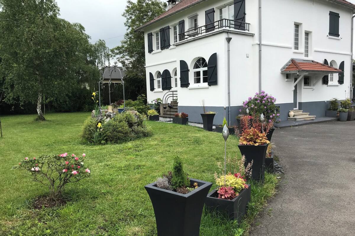 A white building with black shutters, a balcony, and a garden with various plants and flowers.