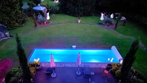 A rectangular swimming pool with a tiled edge, surrounded by a grassy lawn and a few trees. There are two umbrellas and a few lounge chairs near the pool. - Régine MAUFFREY (Champagney)