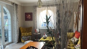 A living room with a sofa, a coffee table, a decorative plant, and a window with curtains. - Régine MAUFFREY (Champagney)