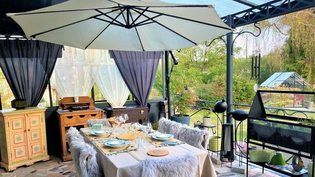 An outdoor patio with a table set for dining, surrounded by a large umbrella, a wicker chest, and a decorative cabinet. - Régine MAUFFREY (Champagney)