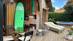 A wooden deck with a glass table, wicker chairs, a lounge chair, and a surfboard. - Wooden house, côte landaise 5min ocean, lakes, ideal family, animals accepted! (Biscarrosse)