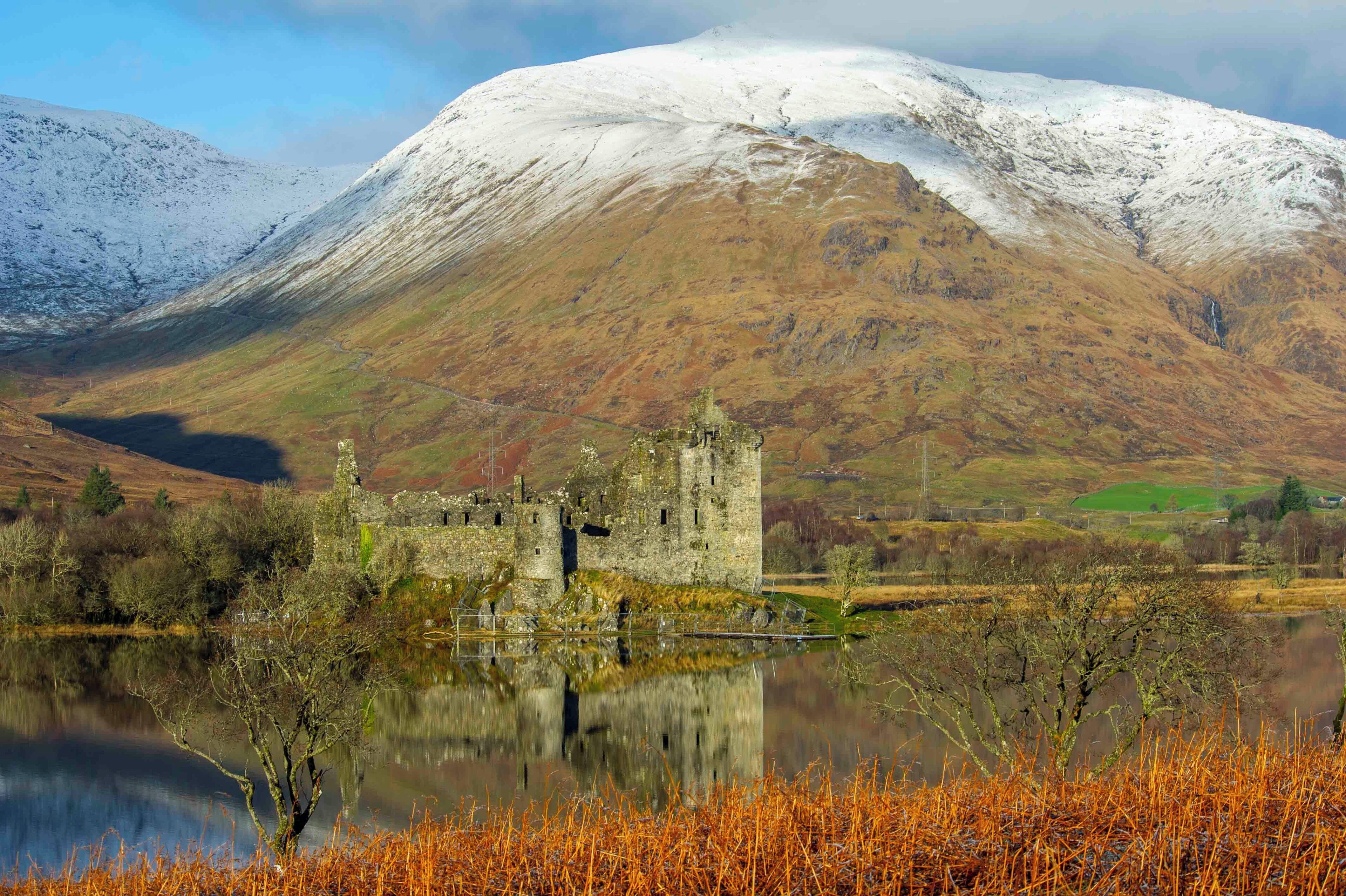 A historic castle by a lake with a snow-capped mountain in the background.