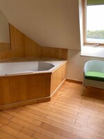 A room with a wooden bathtub, a window with a view, and a wicker chair.