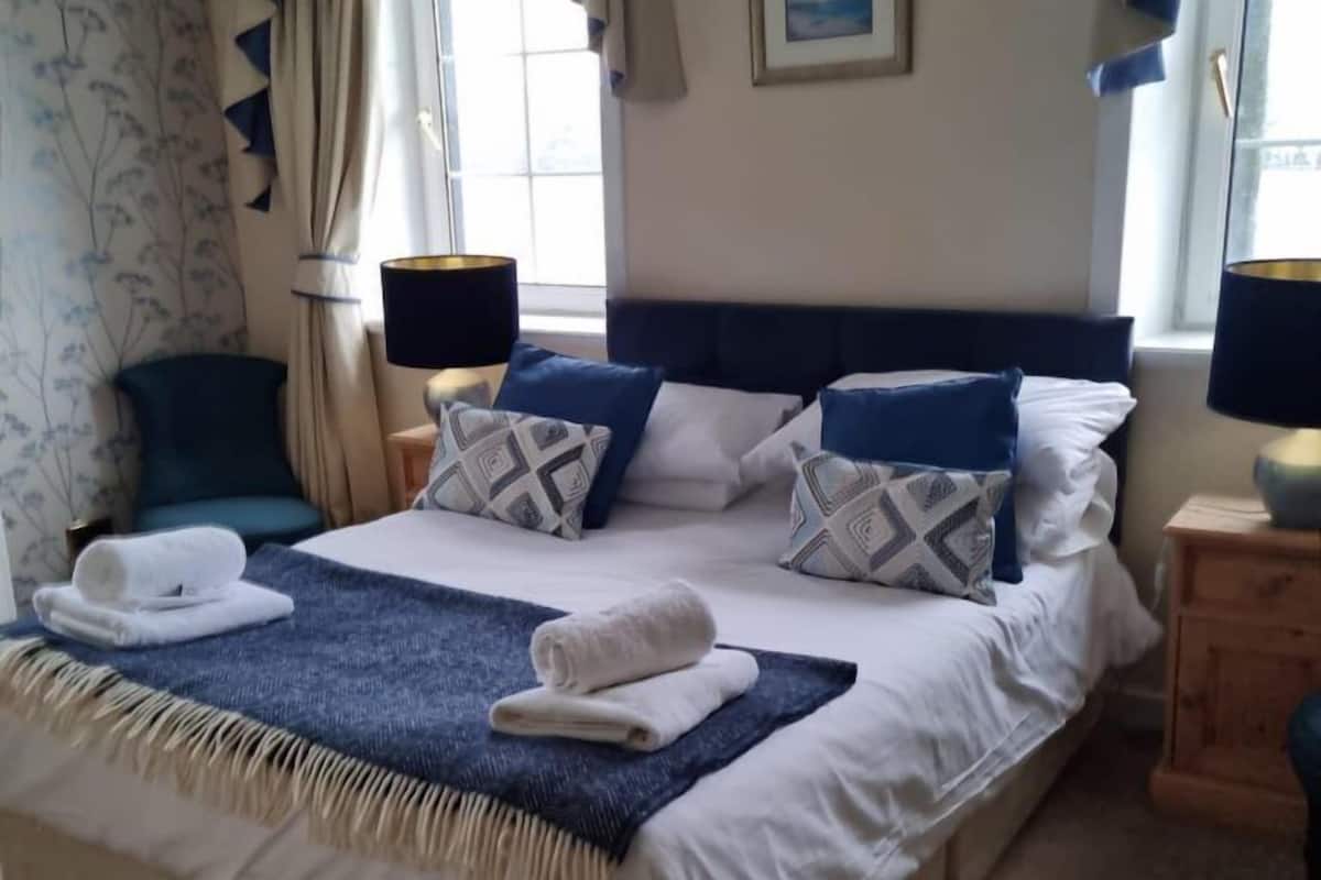 Un lit bien fait, avec une couverture bleue et des oreillers, une table de chevet avec une lampe, et une fenêtre avec des rideaux.