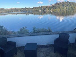 A lakeside patio with two wicker chairs and a small table, overlooking a calm lake and forested hills.