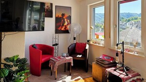 Ein Zimmer mit Fernseher, zwei Stühlen, einem Tisch, einem Ventilator, einem Fenster mit Aussicht und einem Mikrofonständer.