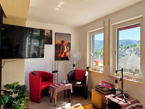 A room with a TV, two chairs, a table, a fan, a window with a view, and a microphone stand. - Helene vacation apartment with balcony and Moselle view at Ferienhaus Moselwelt (Zell (Mosel))
