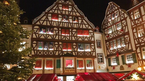 Ein traditionelles Fachwerkhäuschen mit vielen Fenstern, einige mit roten Vorhängen, und ein Weihnachtsbaum mit Lichterketten davor.