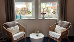A room with two wicker chairs, a white round table, and a view of a residential area through large windows with curtains. - Vacation home "Leuchtturmblick" in the harbor village Rheinsberg (Rheinsberg)